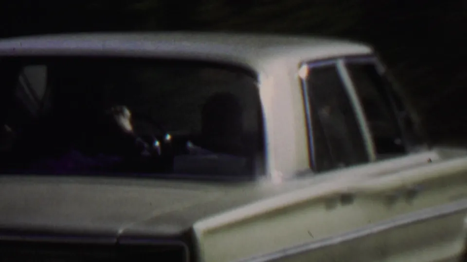 A white 1960s sedan appears close to the camera on a shaded forest road at Farragut State Park, with dark trees behind it and a person faintly visible inside.