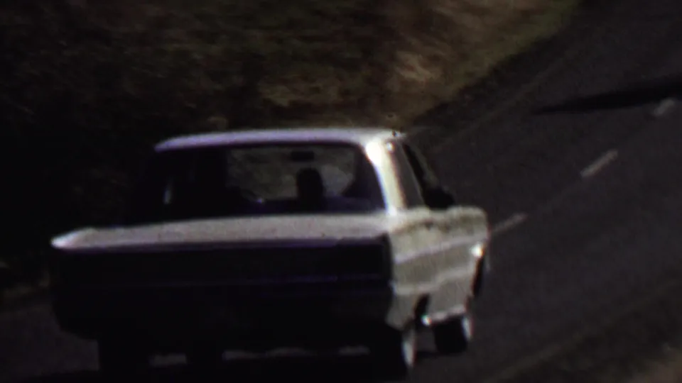 The white sedan drives uphill away from the camera on a curving paved road through the wooded Farragut State Park landscape.