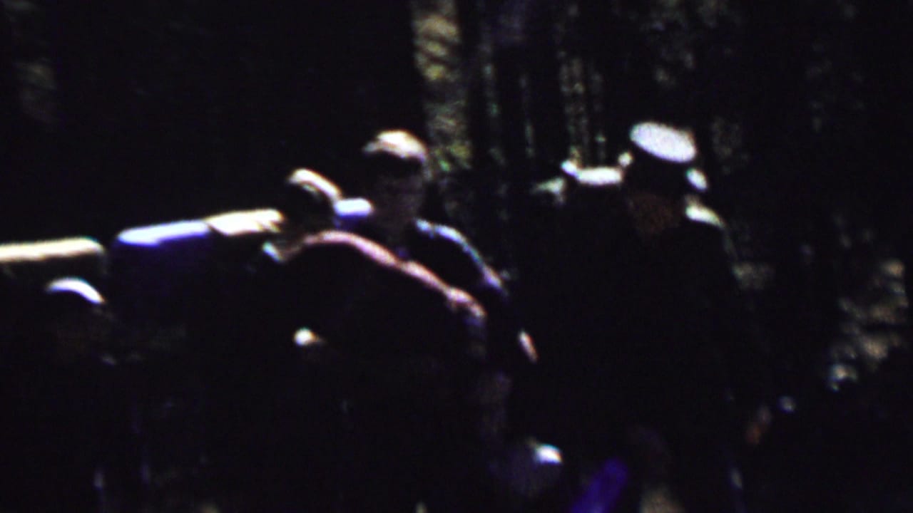 1967 8mm frame from the supplied Farragut State Park clip showing several backpackers in shadowed woods with large packs and dappled light behind them.