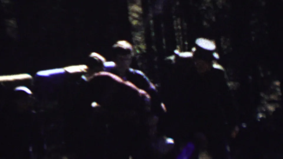 1967 8mm frame from the supplied Farragut State Park clip showing several backpackers in shadowed woods with large packs and dappled light behind them.
