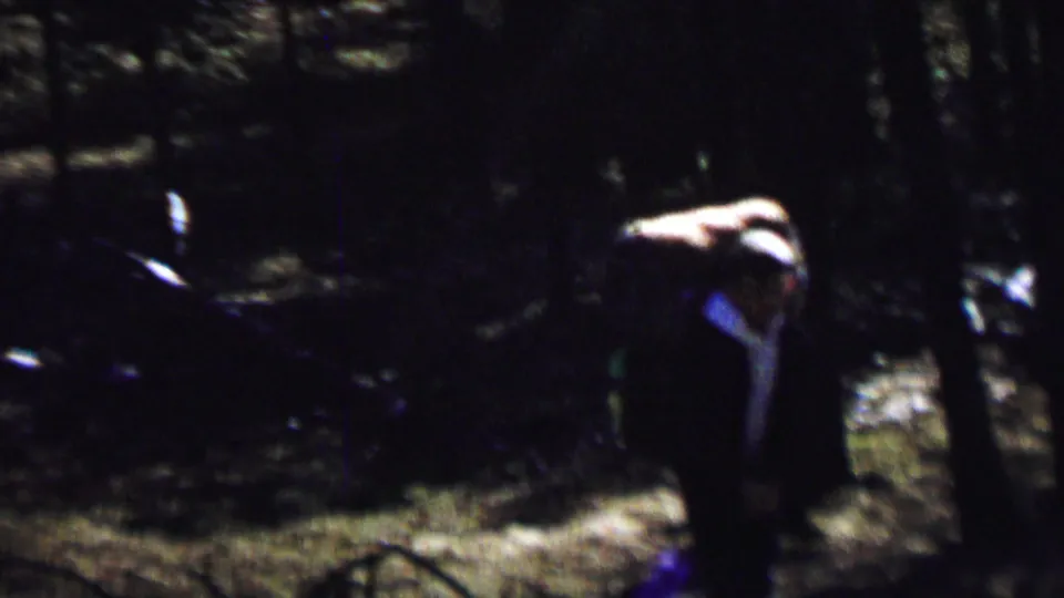 Several hikers are partially visible in a low-light 8mm frame, with a large pack and wooded trail area standing out against the surrounding forest.