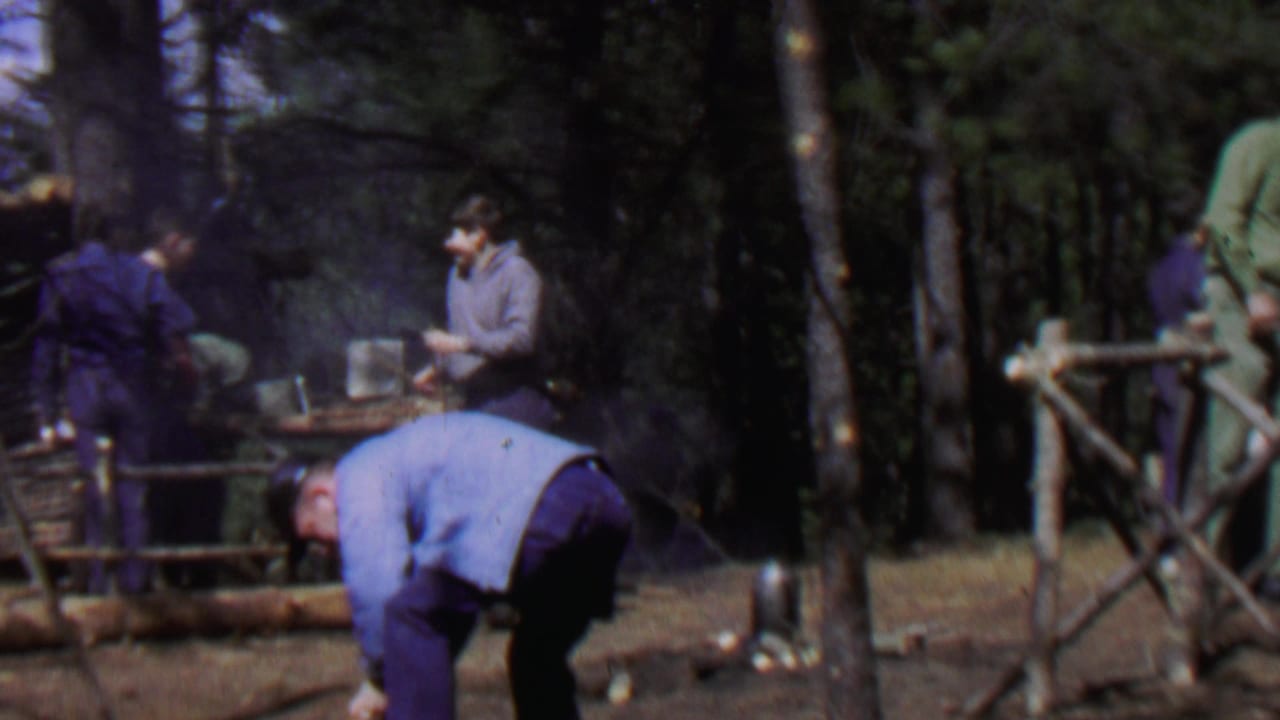 1967 home-movie frame showing men in a forest clearing at Farragut State Park, Idaho, working around cut logs, tools, smoke, and a partly built rustic shelter.