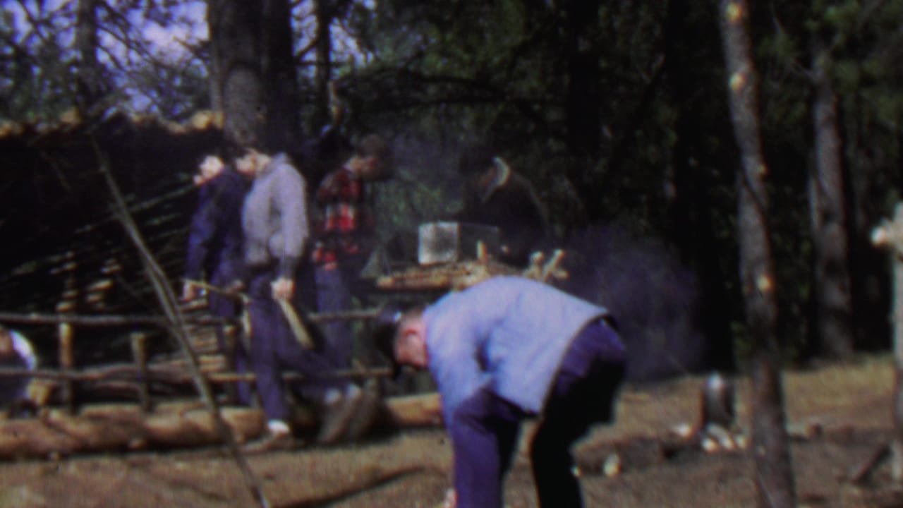 Several men stand near a log shelter frame while a man bends over in the foreground.