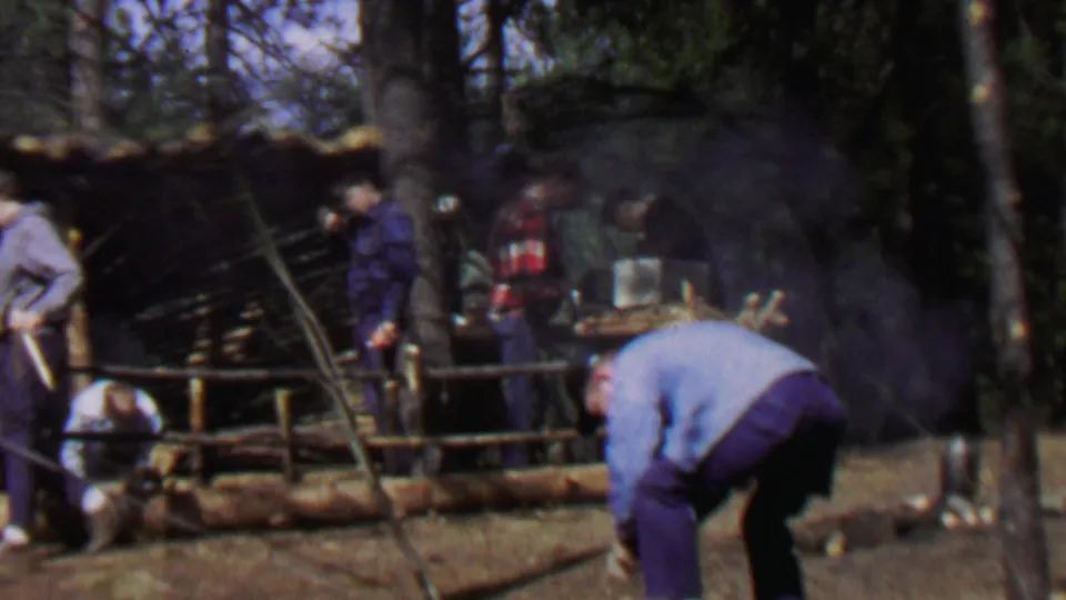 1967 8mm-style frame showing men at a smoky forest work site with cut logs, a rough wooden shelter, and outdoor tools visible in the clearing.