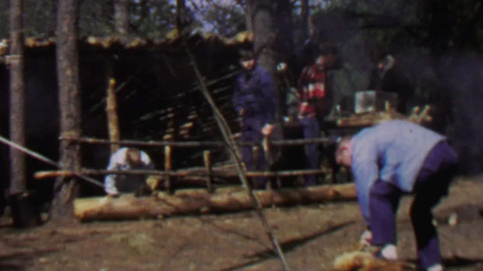 1967 home-movie frame from Farragut State Park showing a rustic log-and-branch shelter, men in the background, smoke, and a foreground man working near a cut log.