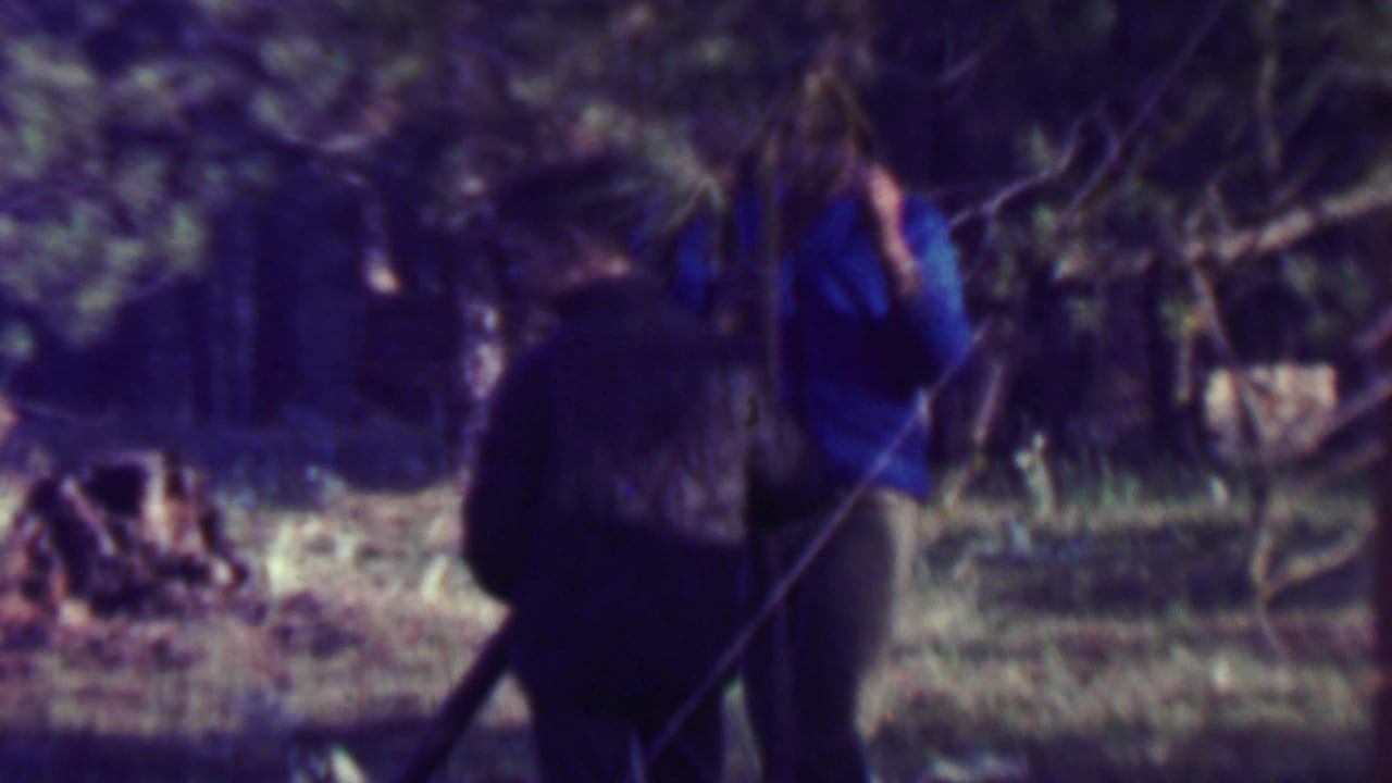 Two adult men at a wooded campsite in supported 1967 Farragut State Park home-movie footage, with one figure in a blue shirt and another darker figure nearby.