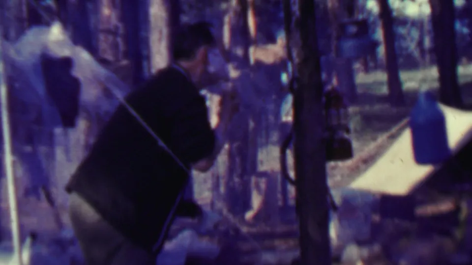 An adult man in a dark top stands near tent fabric, hanging gear, and trees in the supported 1967 Farragut State Park campsite footage.