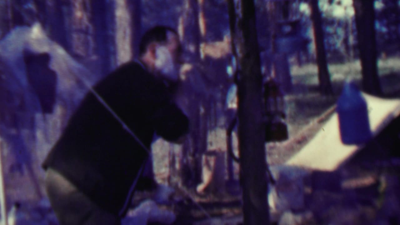 In 1967 Farragut State Park footage, a man in a dark sweater bends beside a forest campsite with tent fabric, trees, and hanging camp gear visible.