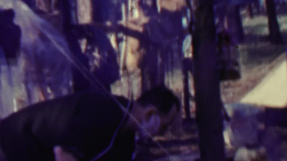 The man bends low in the foreground of a forest campsite, with pale tent fabric, trees, and campsite equipment visible through the purple-blue 8mm color cast.