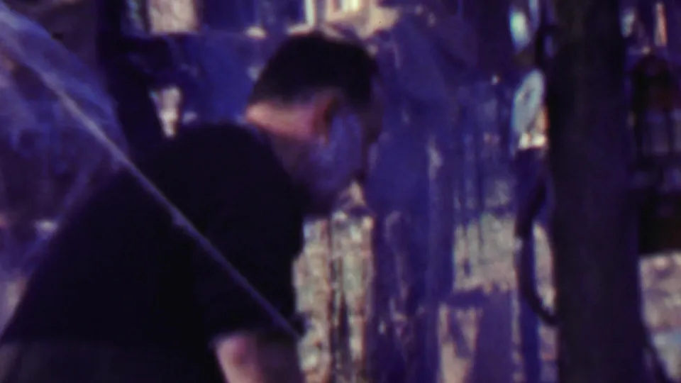 A closer handheld frame shows the man bending beside tree trunks at the campsite, with tent material and dark camp gear partly visible in the background.