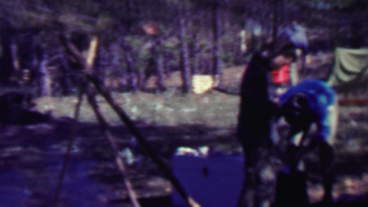 Two adults at a forest campsite in 1967 Farragut State Park footage stand near a canvas tent, boxes, and a tripod-like support.