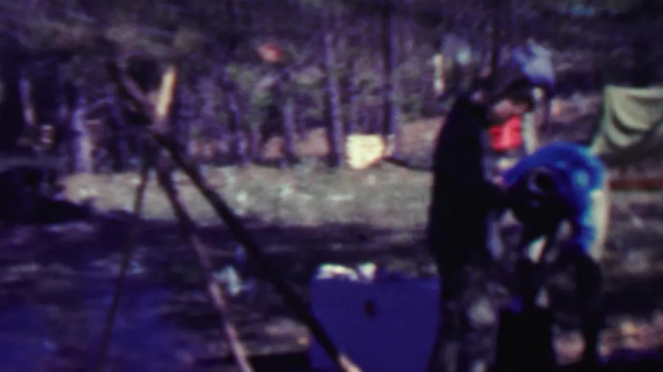Two adults at a forest campsite in 1967 Farragut State Park footage stand near a canvas tent, boxes, and a tripod-like support.