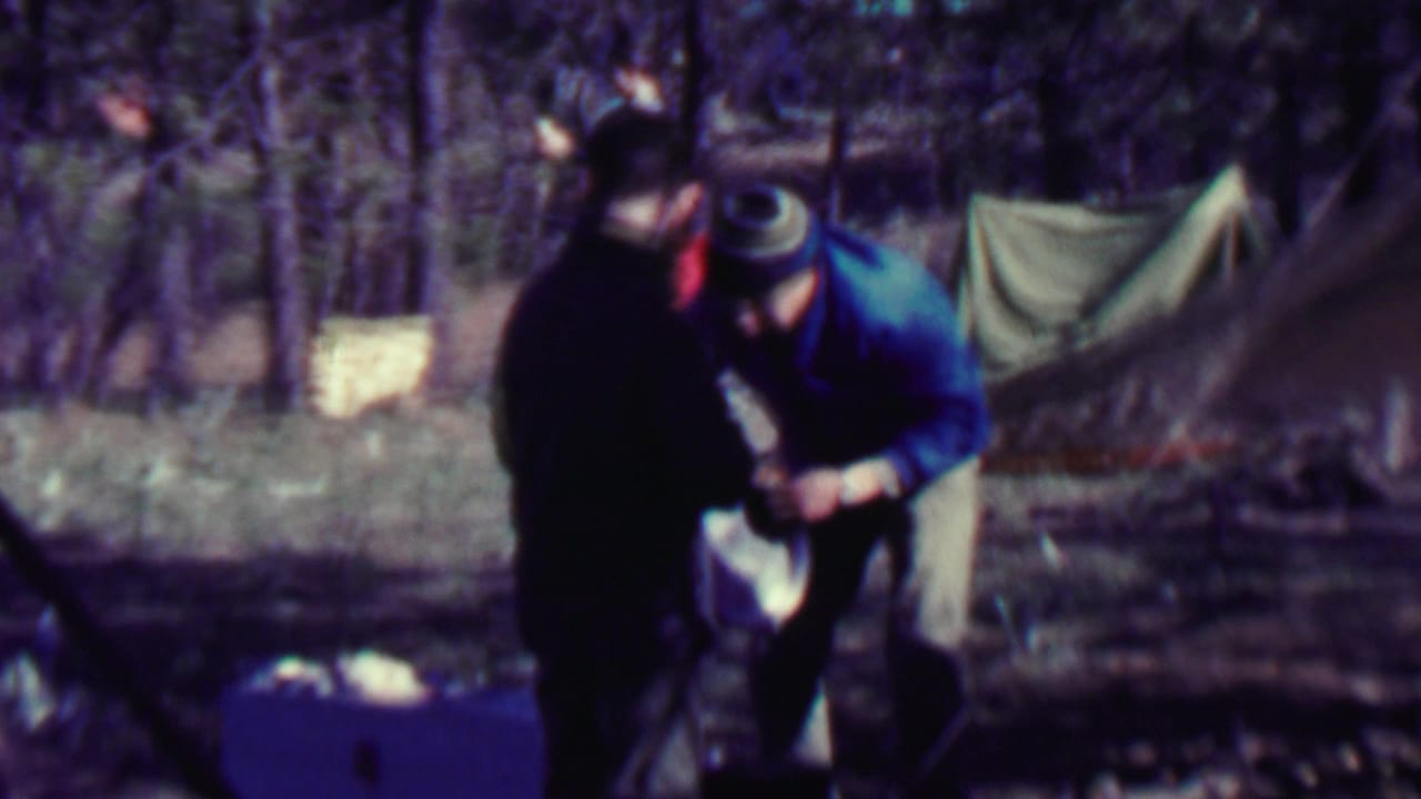 Two people lean over camping gear beside a tent in the forest.