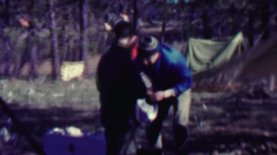 Two adults in outdoor clothing appear to handle or inspect gear beside a canvas tent in wooded 1967 Farragut State Park home-movie footage.