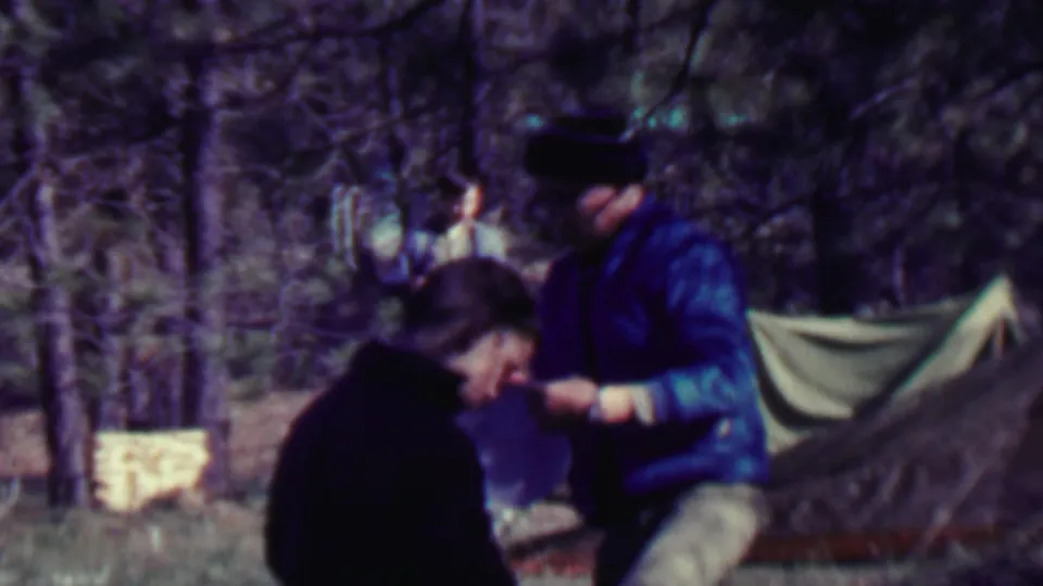 At the forest campsite, an adult in a blue jacket and dark hat bends or sits near another adult with a canvas tent visible behind them.