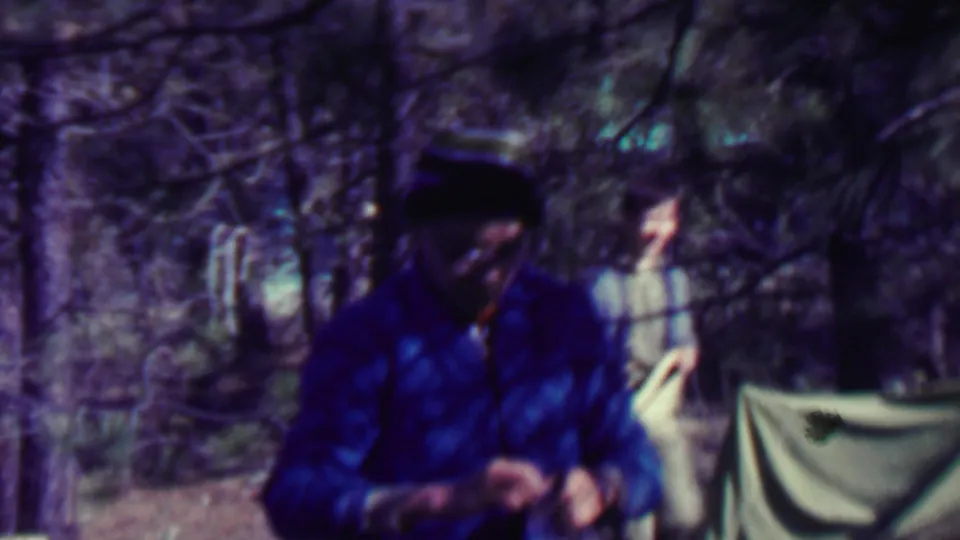 A person in a blue jacket and dark hat stands at a wooded campsite with a canvas tent or tarp and pale hanging object behind them.