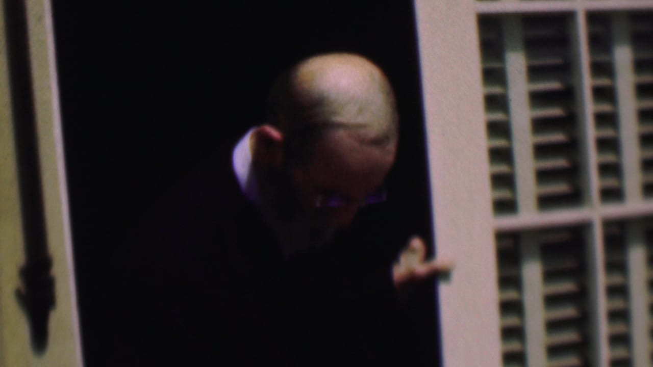 In 1961 San Diego home-movie footage, an older bearded man with eyeglasses and a dark suit stands partly in shadow at a doorway with white louvered shutters.