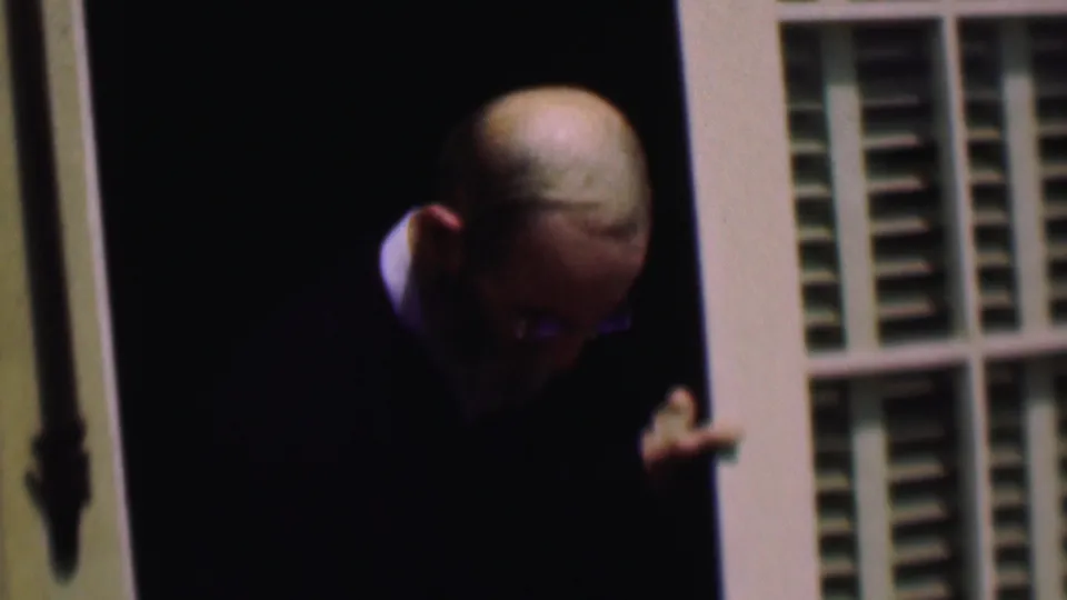 In 1961 San Diego home-movie footage, an older bearded man with eyeglasses and a dark suit stands partly in shadow at a doorway with white louvered shutters.