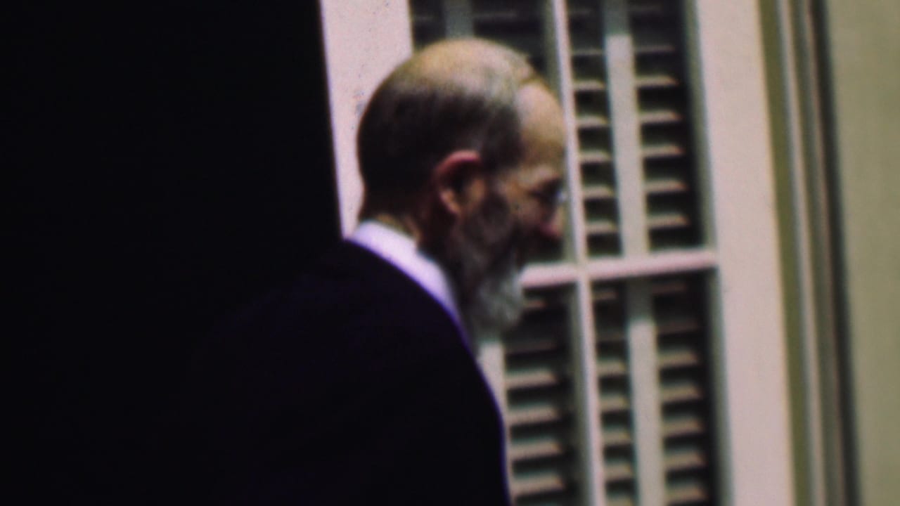 Older man in a dark suit walks past a doorway and shuttered window in profile.