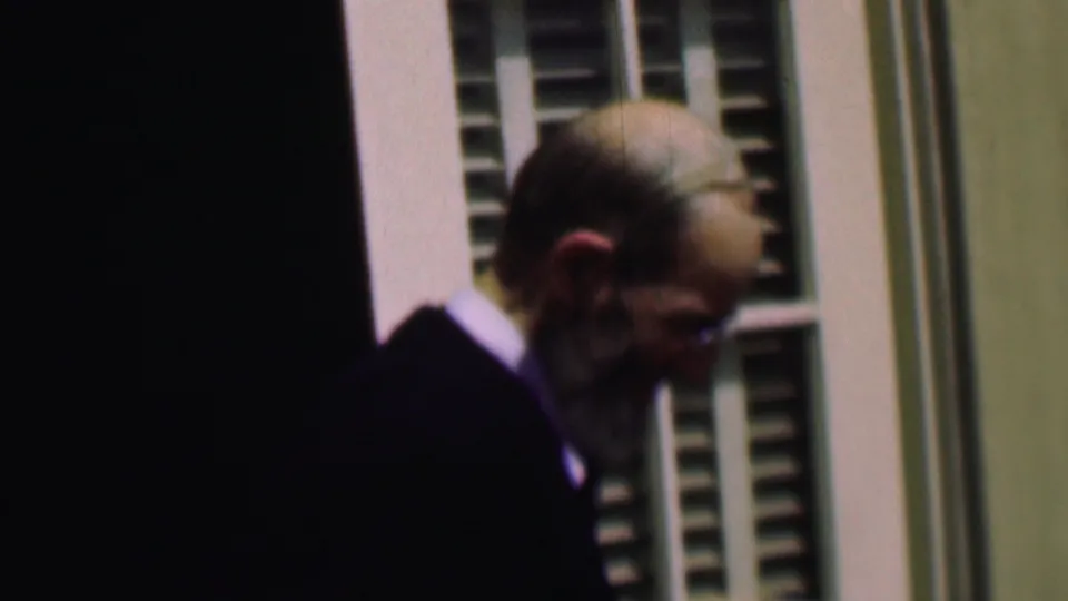 The older man with eyeglasses and a full beard looks downward as he moves beside the doorway and white shutters in soft 8mm color.