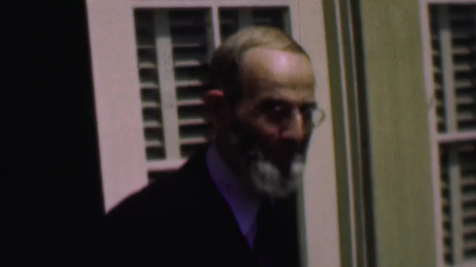 Near the doorway and shuttered window, the older bearded man in a dark suit turns partly toward the camera in the 1961 San Diego home-movie footage.