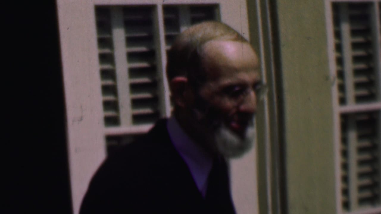 Older bearded man with glasses and a dark suit appears beside white louvered shutters in 1961 San Diego home-movie footage.