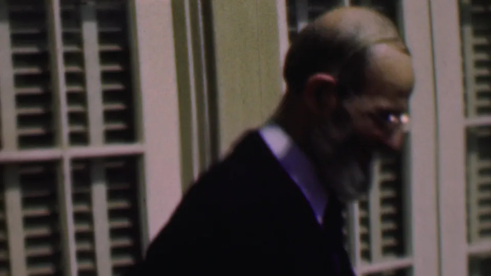 The older gentleman is seen closer in profile, with glasses catching light as he leans or turns beside the shuttered window area.
