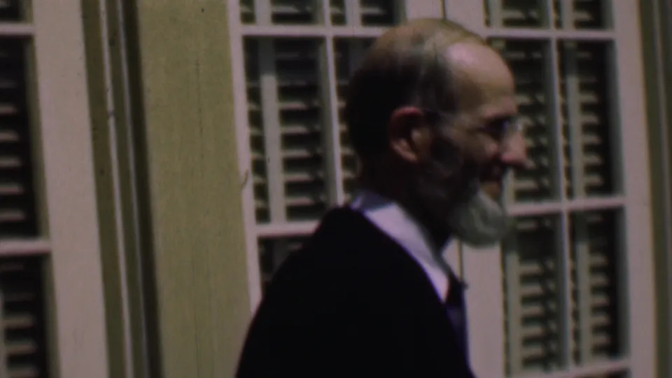 The suited older man smiles in profile beside white louvered shutters, suggesting a friendly greeting or response in the 1961 home-movie clip.