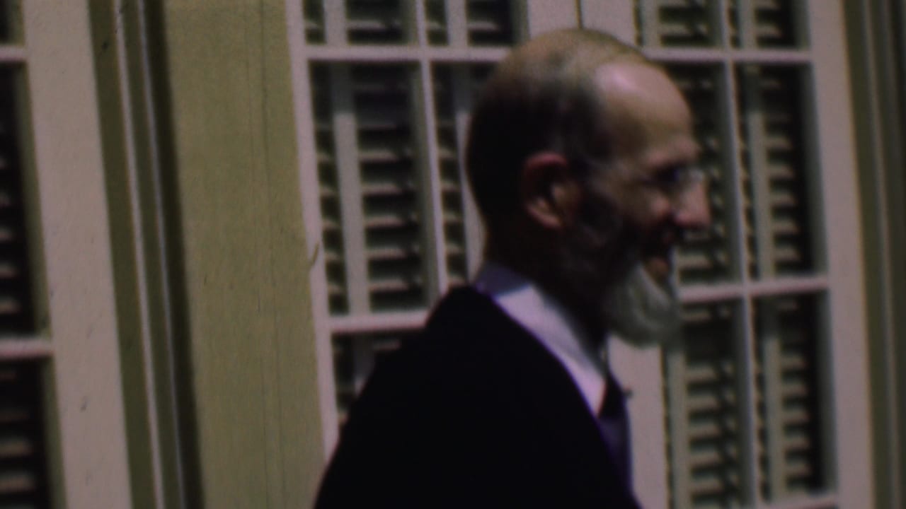 In supplied 1961 home-movie footage from San Diego, a bearded adult man wearing glasses, a dark jacket, light shirt, and purple tie walks in profile past white-framed windows with green shutters.
