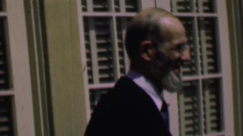 In supplied 1961 home-movie footage from San Diego, a bearded adult man wearing glasses, a dark jacket, light shirt, and purple tie walks in profile past white-framed windows with green shutters.