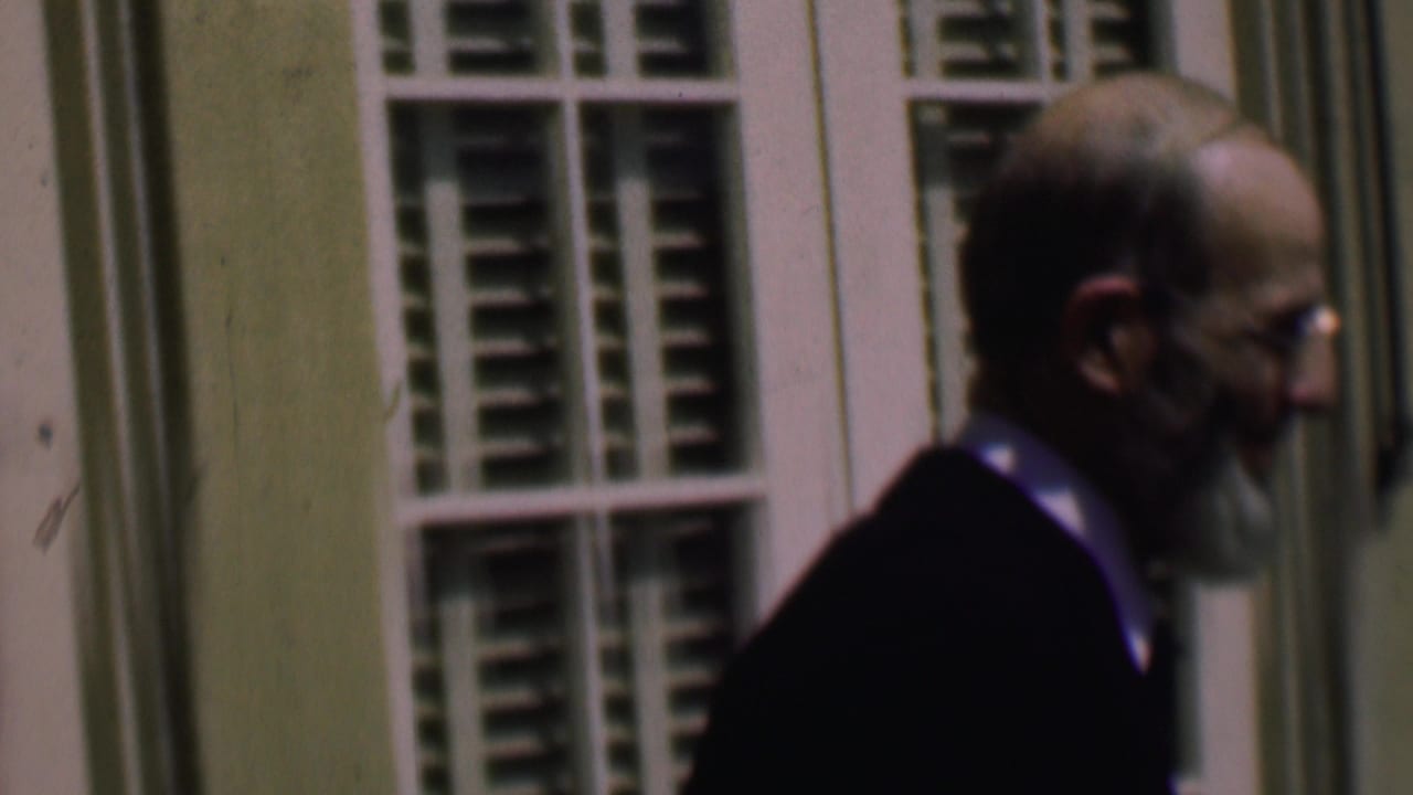 The bearded man remains in side profile as he moves along the exterior wall, with shuttered windows and pale trim behind him in the 8mm frame.