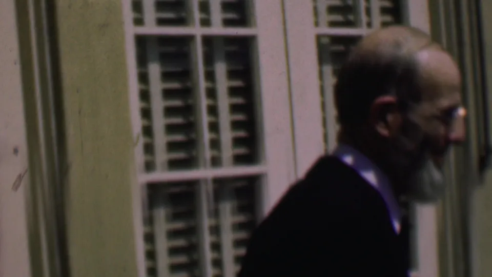 The bearded man remains in side profile as he moves along the exterior wall, with shuttered windows and pale trim behind him in the 8mm frame.