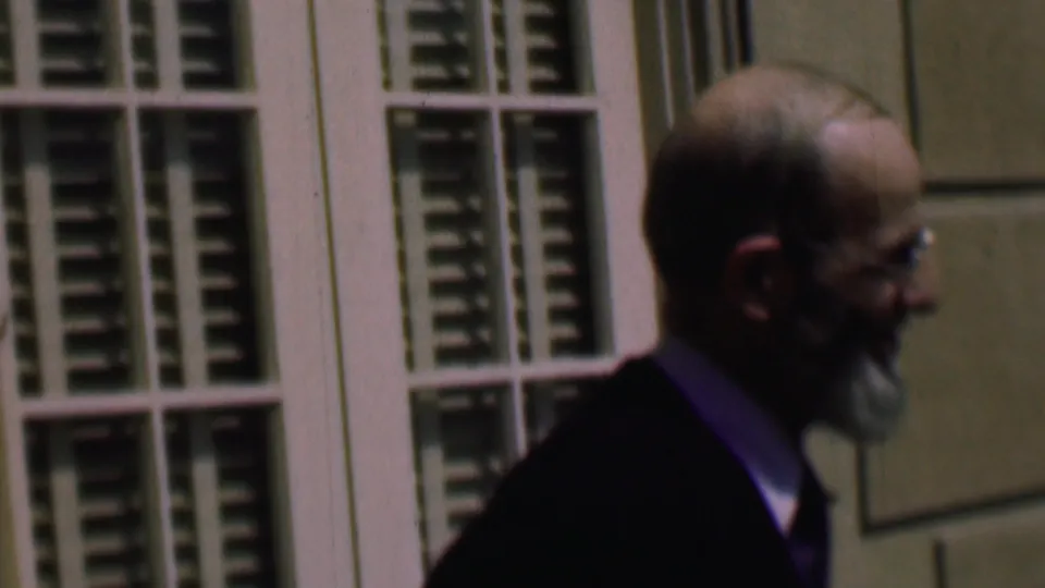 A closer side-profile view shows the man with glasses and beard walking past white-framed windows, with soft focus, grain, and muted color visible in the 8mm scan.