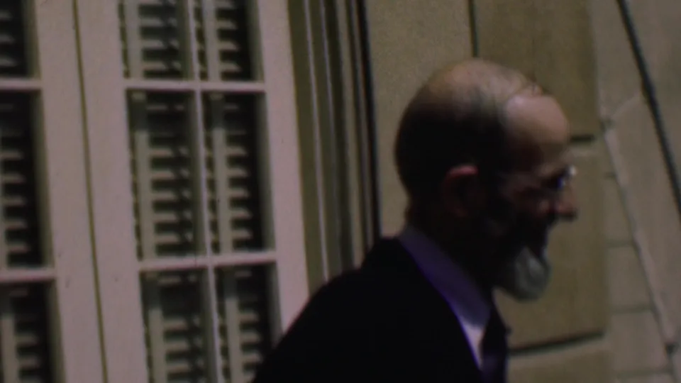 Near the end of the clip, the suited man continues past the green shutters and exterior wall panels, partly blurred by motion and handheld 8mm capture.