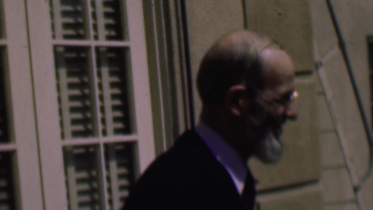 Adult man in a dark suit and glasses walks past a pale exterior building with white louvered windows in the supplied 1961 San Diego home-movie clip.