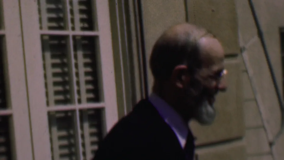 Adult man in a dark suit and glasses walks past a pale exterior building with white louvered windows in the supplied 1961 San Diego home-movie clip.