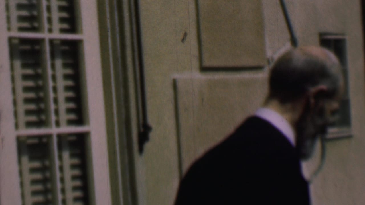 Dark-suited man seen in profile near a shuttered window on a pale building.