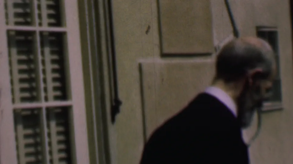 The suited man continues across the frame in profile beside a pale building facade and shuttered or louvered window, with handheld 8mm softness visible.