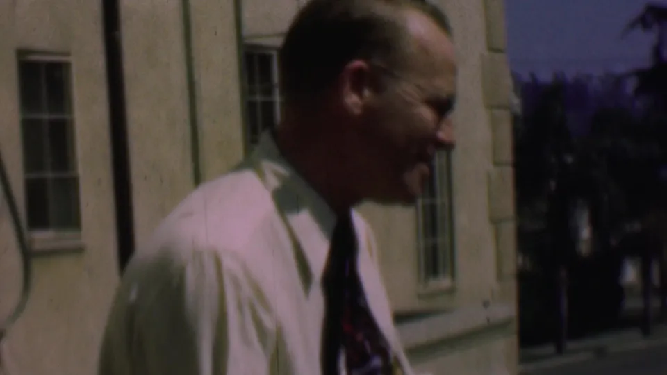 A second adult man in a light shirt and patterned tie walks past the same pale exterior building, with a window and trees visible behind him.