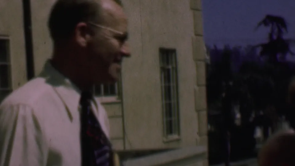 The man in a light shirt and tie remains in profile near the building corner, with louvered or multi-pane windows and dark trees in the background.