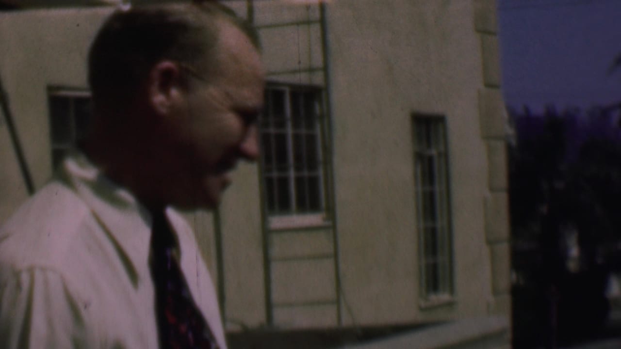 In supported 1961 San Diego home-movie footage, an adult man in a light shirt and patterned tie walks close to the camera beside a building with barred windows.
