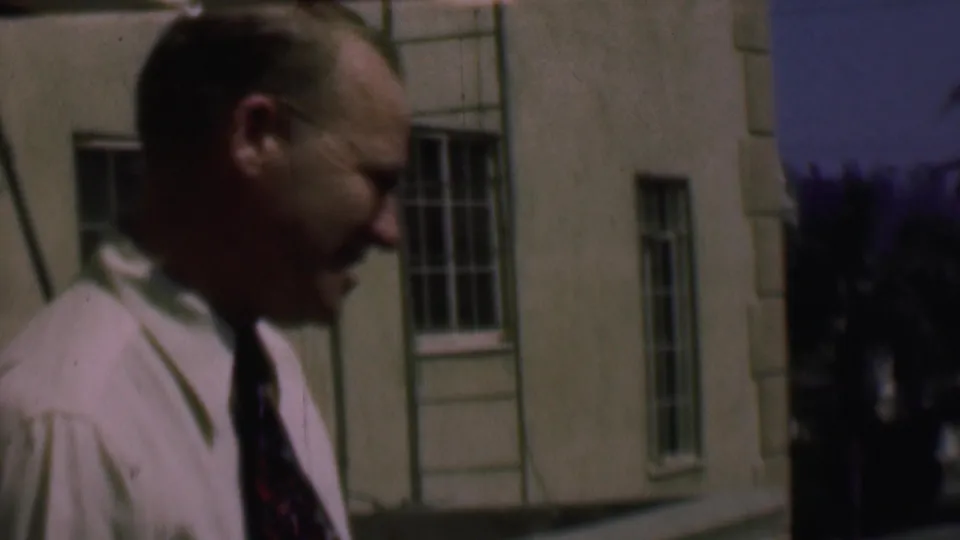 In supported 1961 San Diego home-movie footage, an adult man in a light shirt and patterned tie walks close to the camera beside a building with barred windows.