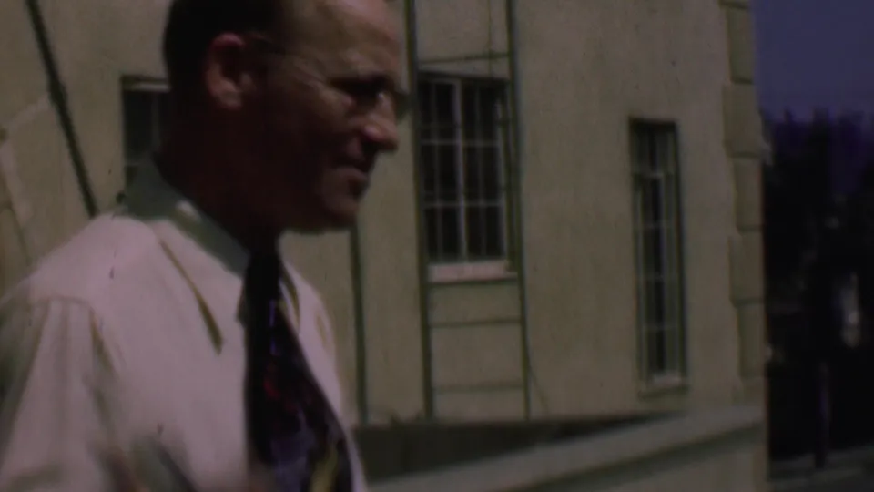 The 1961 San Diego home-movie frame shows a close side view of an adult man in a light dress shirt and tie passing a pale building exterior with barred windows.