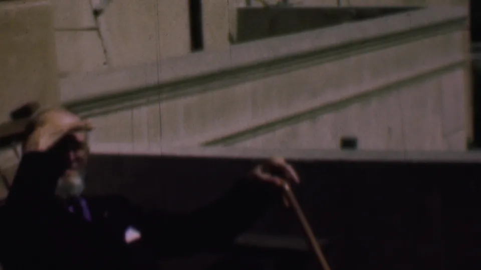 An older bearded man in a dark suit appears in shadow while holding a wooden cane near a sunlit exterior wall in the supported 1961 San Diego home-movie scene.