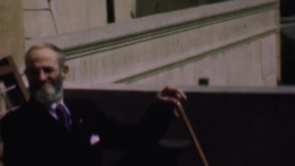 The older bearded man stands or poses with a wooden cane, facing the camera from a shaded area beside a sunlit building exterior in the 1961 San Diego footage.