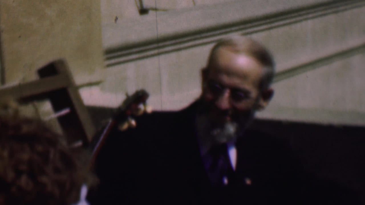 In this 1961 home-movie frame from supplied San Diego metadata, an older man in a dark suit and glasses sits near a stringed instrument while another person is partly cropped at the left edge.