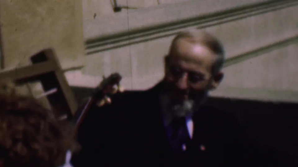In this 1961 home-movie frame from supplied San Diego metadata, an older man in a dark suit and glasses sits near a stringed instrument while another person is partly cropped at the left edge.