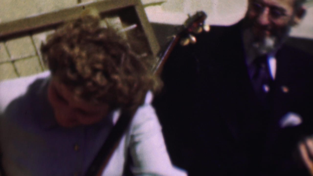 Curly-haired woman leans forward beside a suited man and a stringed instrument.