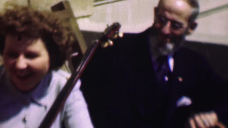 The curly-haired woman smiles at the left of the frame while the older man in a dark suit and glasses sits beside the neck of a stringed instrument in the supplied 1961 home-movie clip.