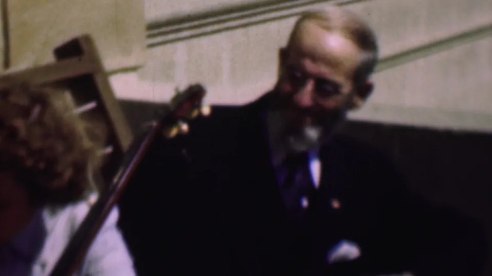 An older man wearing glasses, a dark suit, tie, and pocket square appears behind the neck of a stringed instrument in a softly focused indoor home-movie frame.